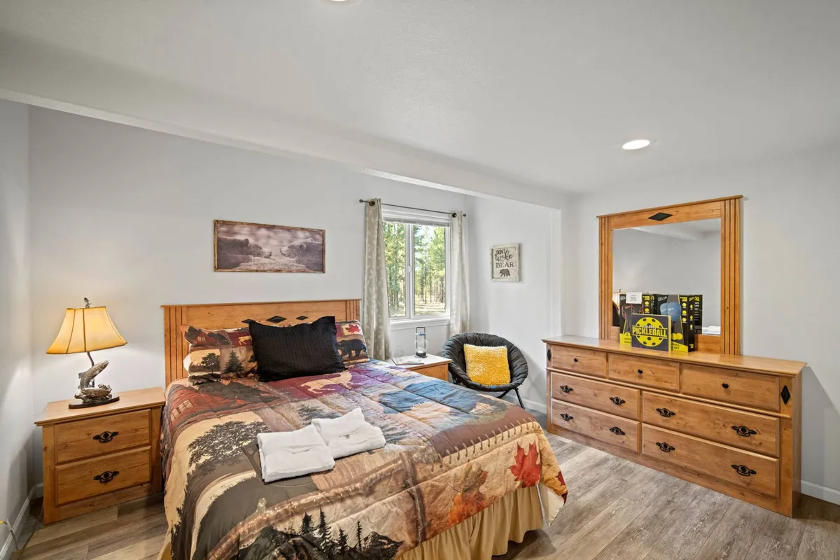A comfortable bedroom with a bed covered by a colorful quilt, a wooden dresser, a large window offering a view, and a black chair. Natural light streams in, enhancing the feeling of peace and privacy.