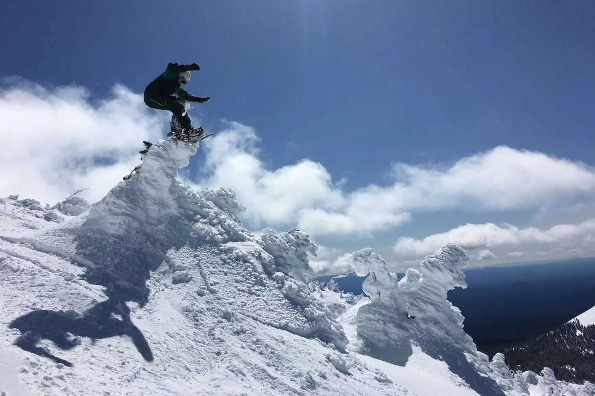 A snowboarder soars above a snow-covered mountain peak, framed by dramatic clouds and expansive blue sky. The image captures the exhilaration of winter sports with panoramic alpine scenery in the background.