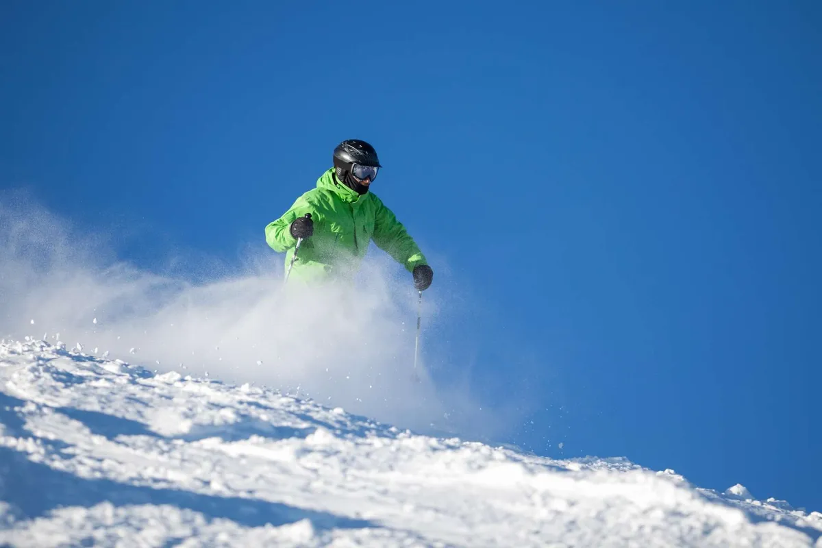 A skier clad in a bright green jacket descends a pristine snowy slope beneath a clear blue sky. The vast, open mountain terrain and vibrant energy evoke the excitement of outdoor exploration in winter.