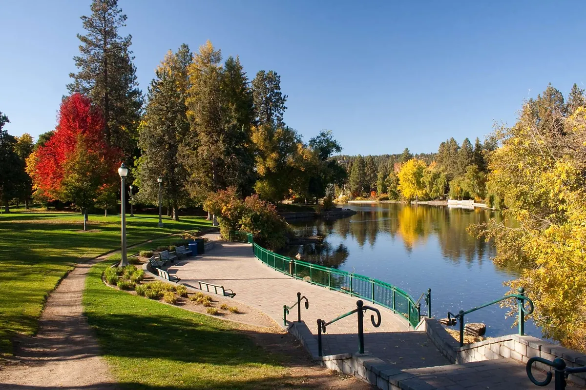 A tranquil park scene with a tree-lined path, reflective water, colorful autumn foliage, and park benches. The green railing and expansive sky add to the sense of peaceful outdoor recreation and community spirit.