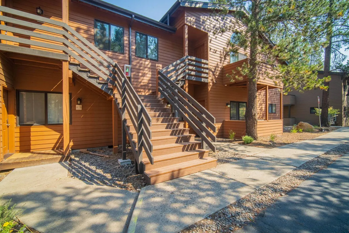 This image captures the exterior of The Fisher Place, highlighting the welcoming wooden staircase, siding, and windows. The scene suggests an inviting environment for guests to gather outdoors and enjoy the natural surroundings, with hints of pine trees visible beyond the building.