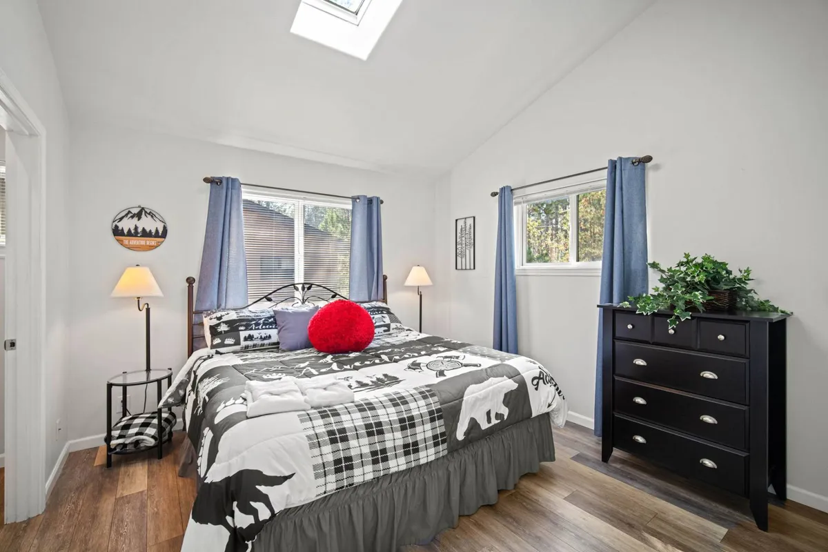 A serene bedroom featuring a bed with patterned bedding, a red accent pillow, a black dresser, and lamps casting a warm light. The atmosphere is calm, inviting rest and relaxation.
