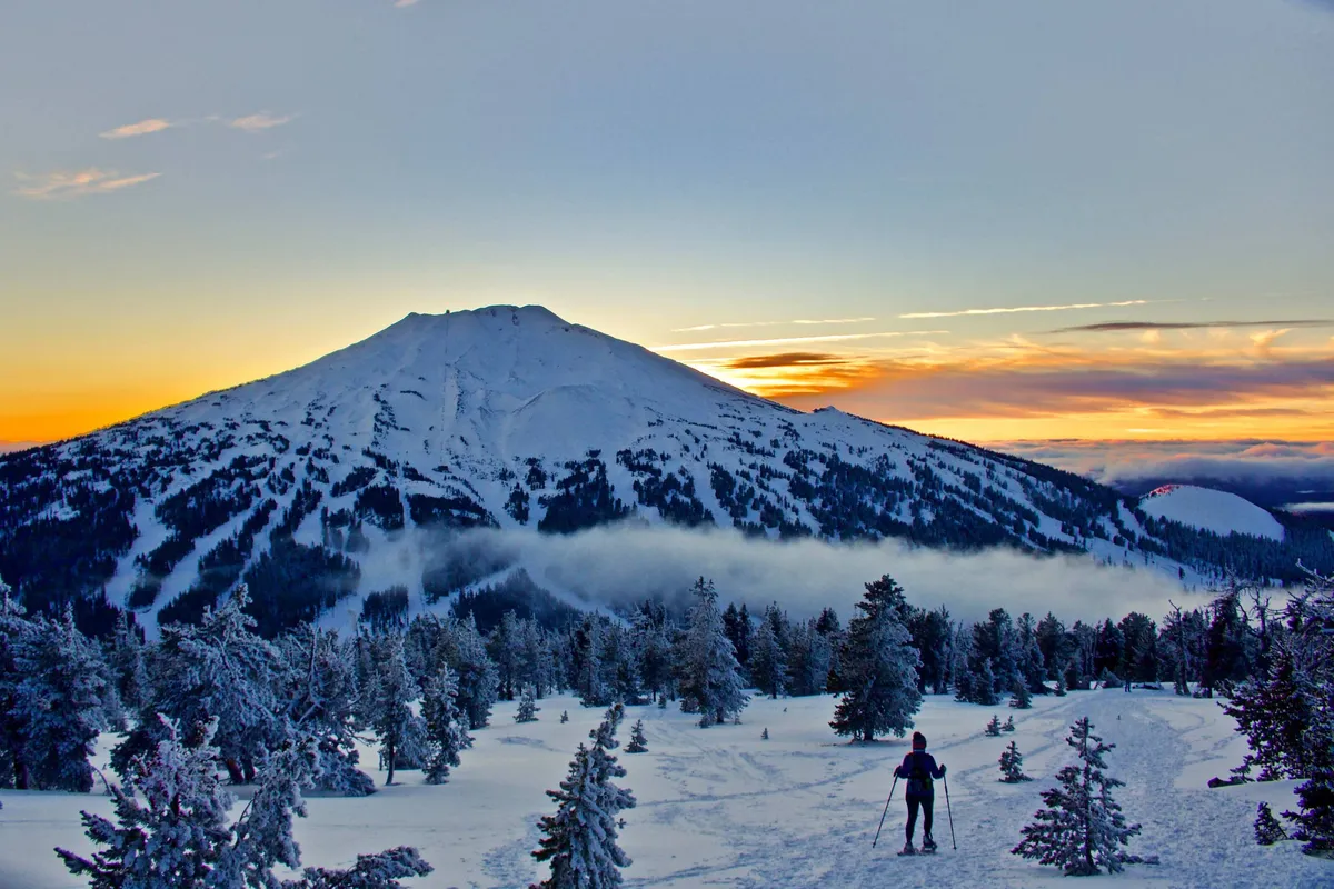 A majestic mountain peak rises above a snow-covered landscape at sunset, creating a serene and awe-inspiring backdrop. The soft, tranquil colors highlight the beauty of the local environment, evoking a sense of adventure and peaceful escapism.