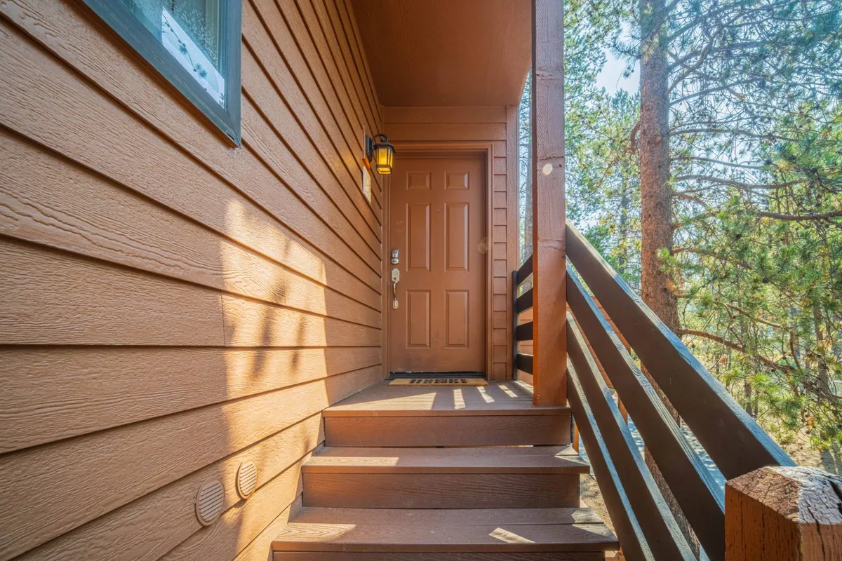 The exterior of The Fisher Place is shown with a welcoming front door, staircase, and outdoor light fixture. The setting is cozy and approachable, with sunlight filtering through the trees, suggesting a safe and comfortable home base where family adventures begin and end.