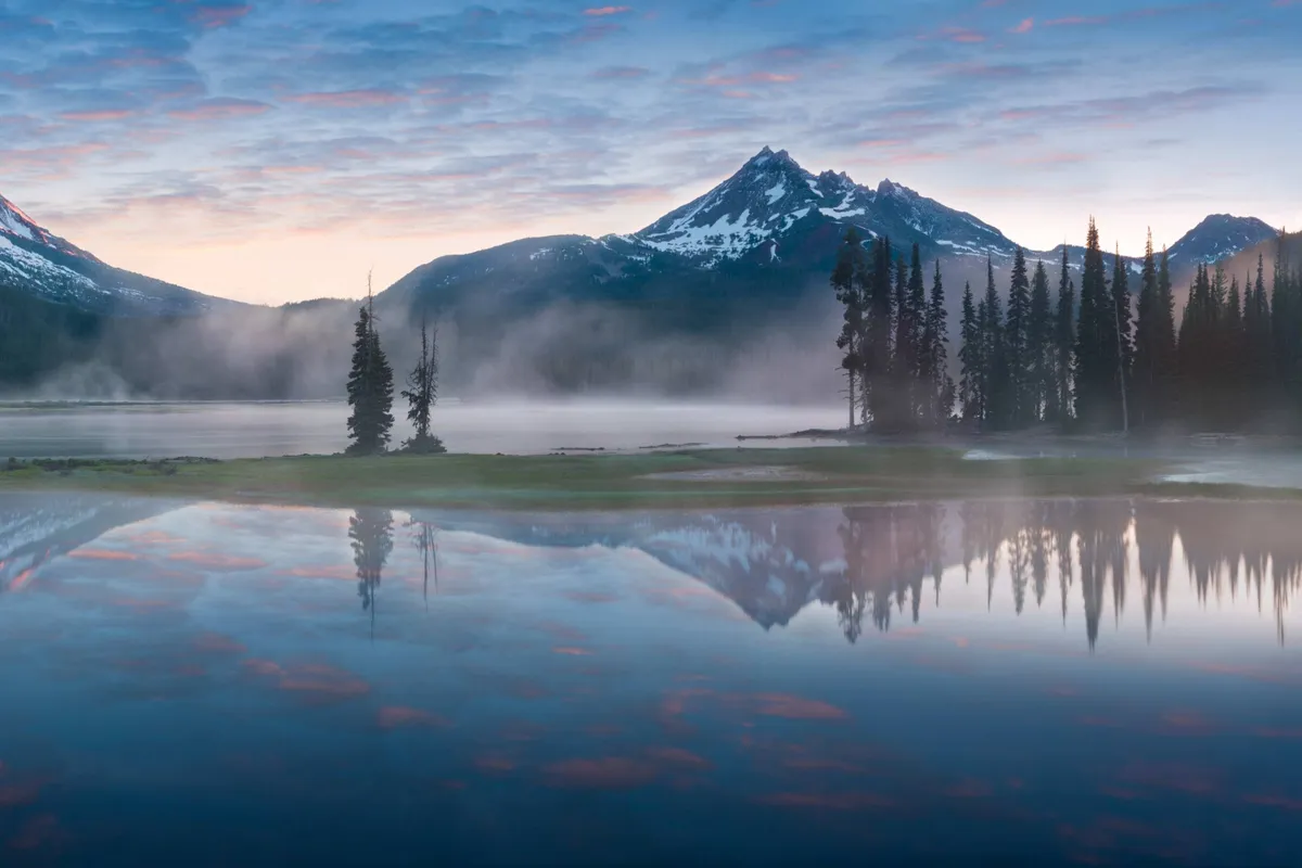 A tranquil view of mountains, a serene lake, and lush trees beneath an open sky. The setting is peaceful and inviting, ideal for outdoor exploration, hiking, and wildlife spotting. The natural beauty emphasizes opportunities for scenic walks, bike rides, or picnics beneath tall pines.